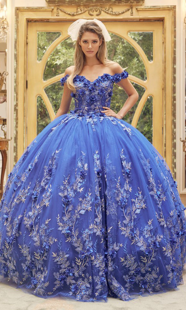 A woman poses elegantly indoors in front of an ornate wooden door, wearing the Juliet 1447 Off-the-Shoulder Quinceañera Dress with beaded embroidery and a small white veil.